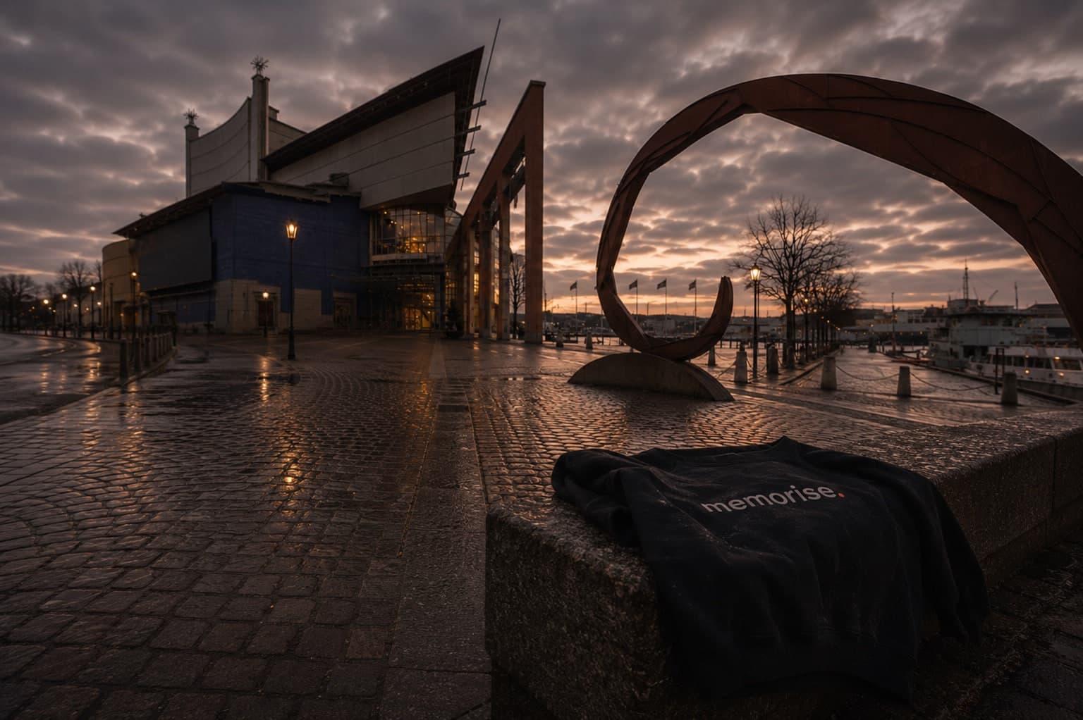 Memorise-hoodie på en stenmur vid Göteborgsoperan i skymning, med en cirkelskulptur i cortenstål, våta kullerstenar och hamnen som speglar solnedgången