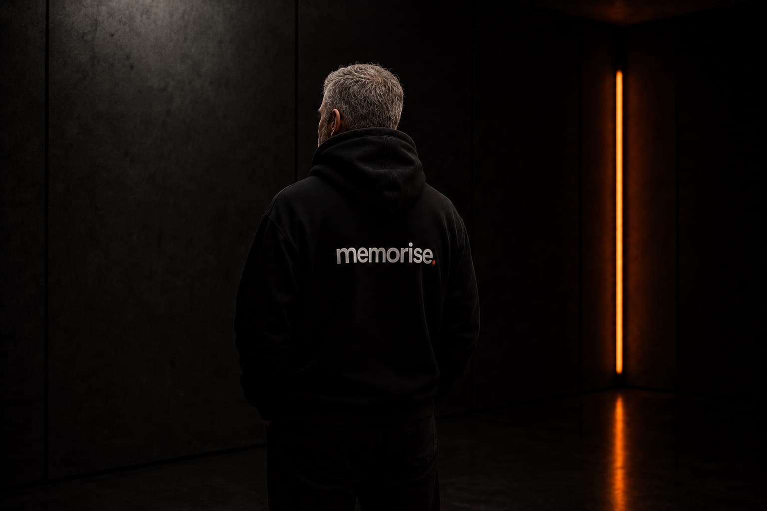 Memorise-specialist sedd bakifrån i hoodie, äldre, kort grått hår, mot mörk vägg med varm orange ljusbar