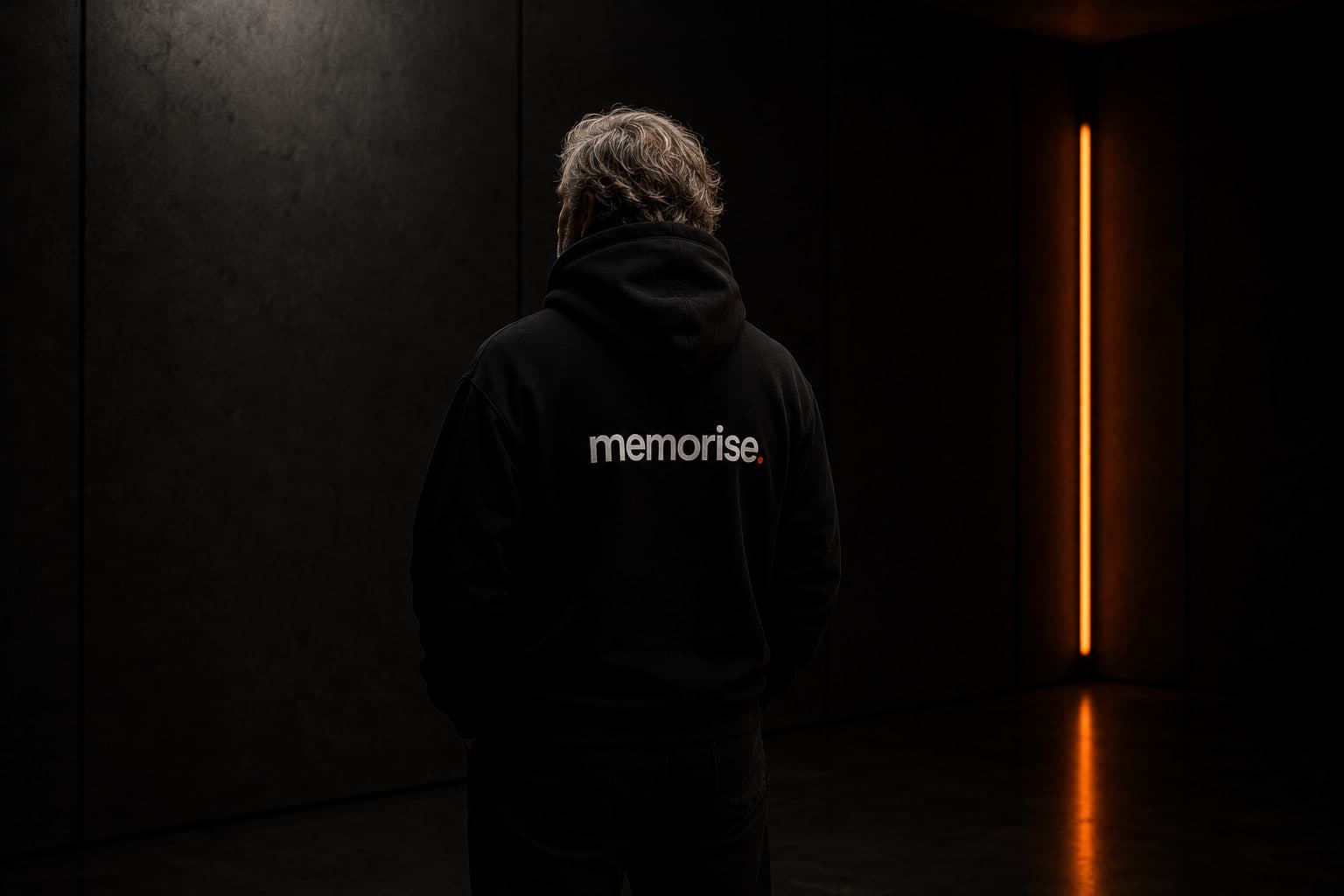 Memorise specialist seen from behind in hoodie, older, wavy hair, against dark wall with warm orange light bar