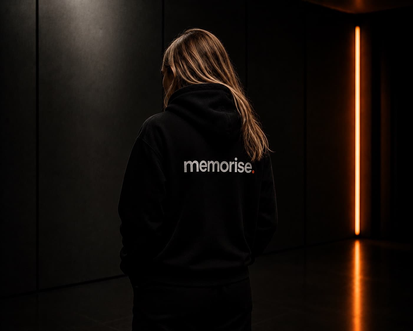 Memorise-specialist sedd bakifrån i hoodie, blont långt hår, mot mörk vägg med varm orange ljusbar