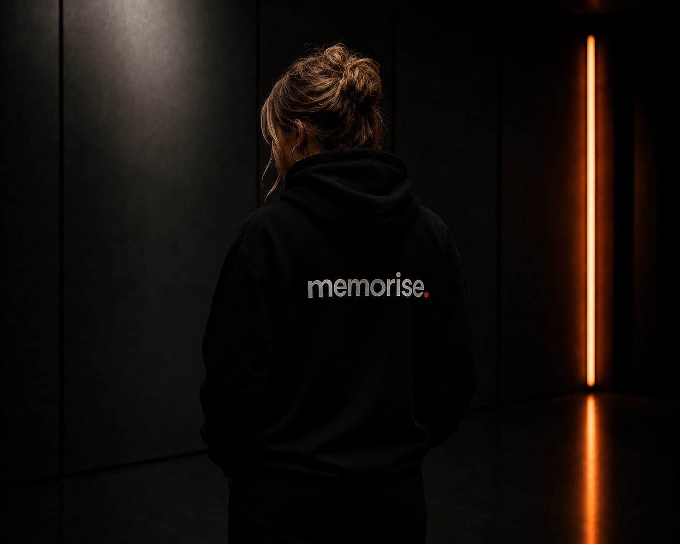 Memorise-specialist sedd bakifrån i hoodie, uppsatt hår, mot mörk vägg med varm orange ljusbar