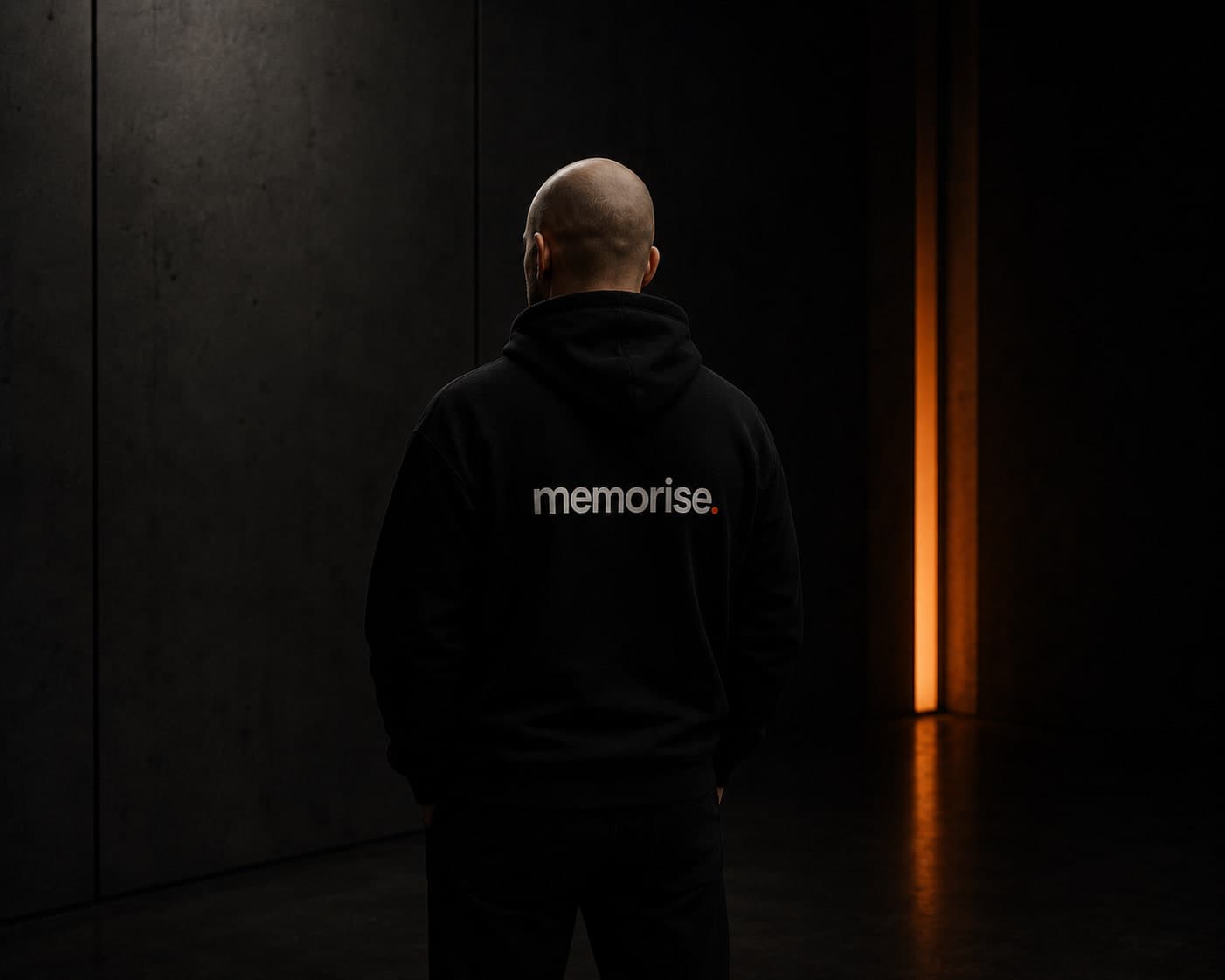 Memorise-specialist sedd bakifrån i hoodie, rakad, mot mörk vägg med varm orange ljusbar