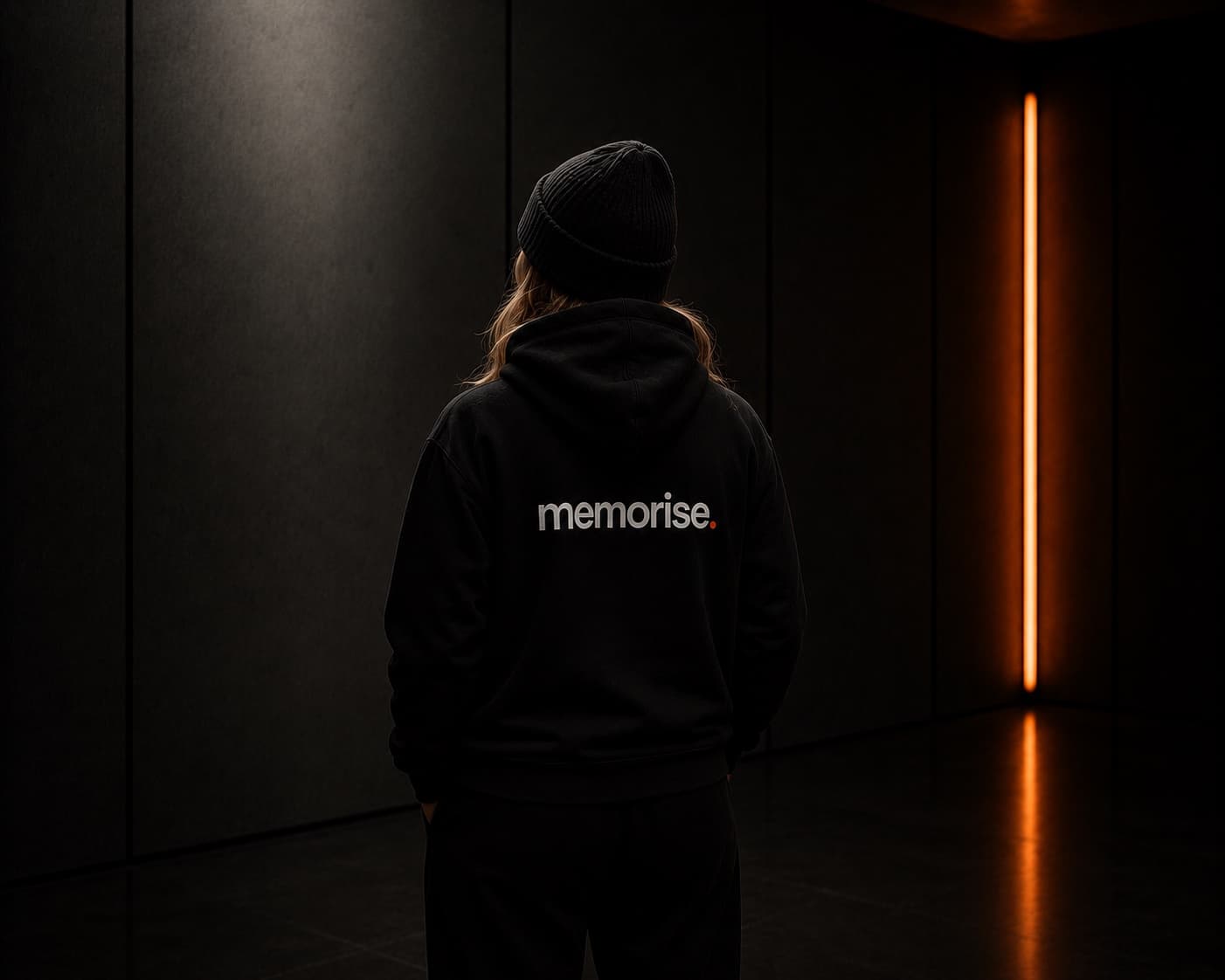 Memorise specialist seen from behind in beanie and hoodie, longer hair, against dark wall with warm orange light bar